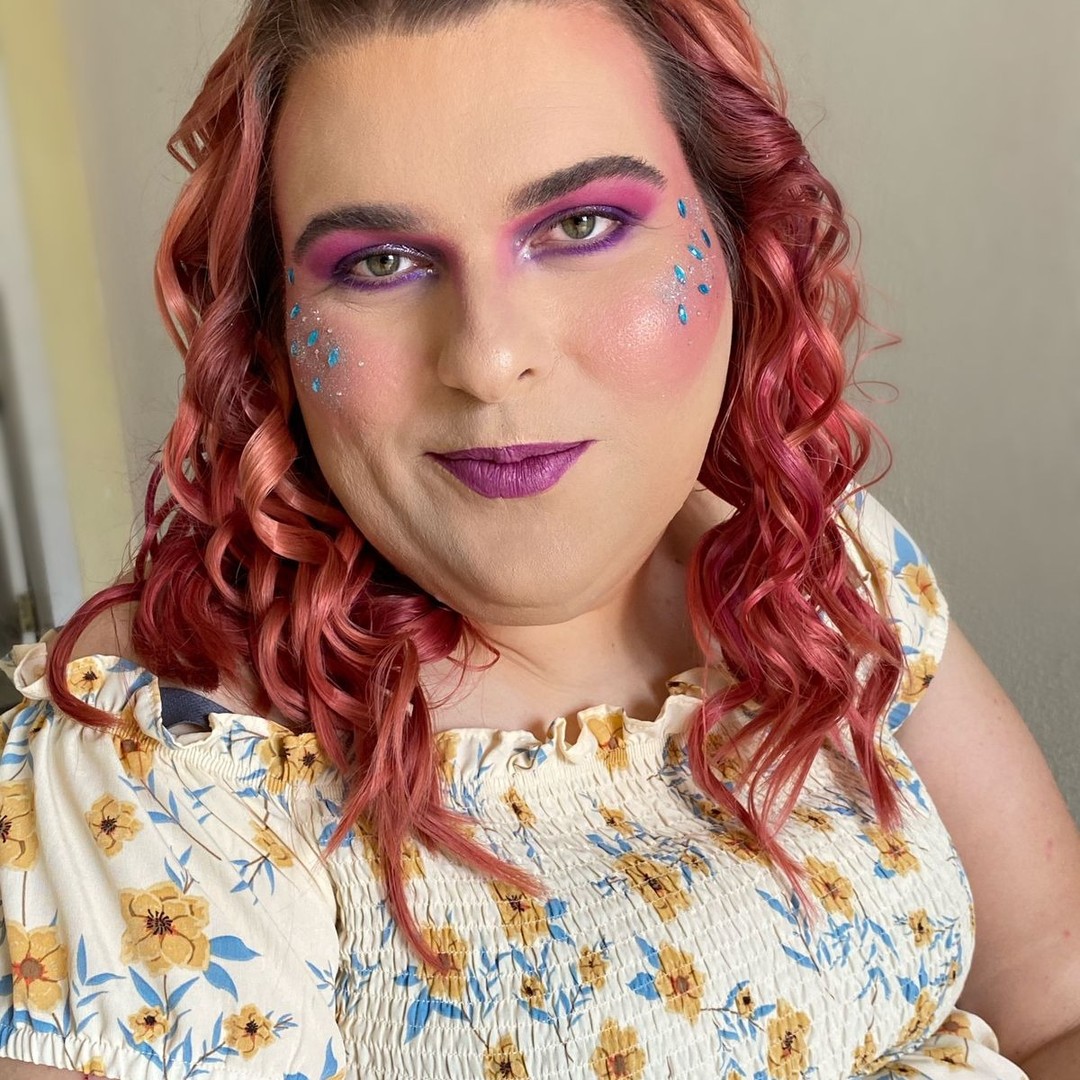 Paloma Macetko sorrindo para a câmera com maquiagem de fada em tons de rosa e roxo, cabelo cacheado rosado